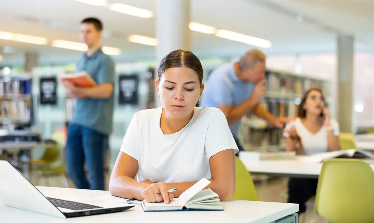 Concentrated young woman checking planner while studying in the library and using laptop
